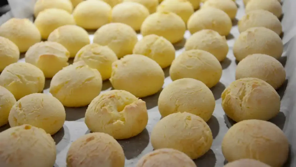 Bandeja com vários pães de queijo com creme de leite, dourados e recém-assados, dispostos sobre papel manteiga.