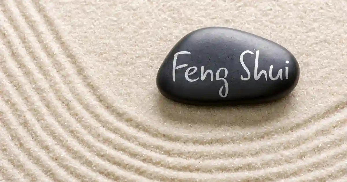 Pedra preta com a inscrição "Feng Shui" sobre areia com círculos concêntricos, simbolizando harmonia e conexão com plantas que atraem prosperidade.