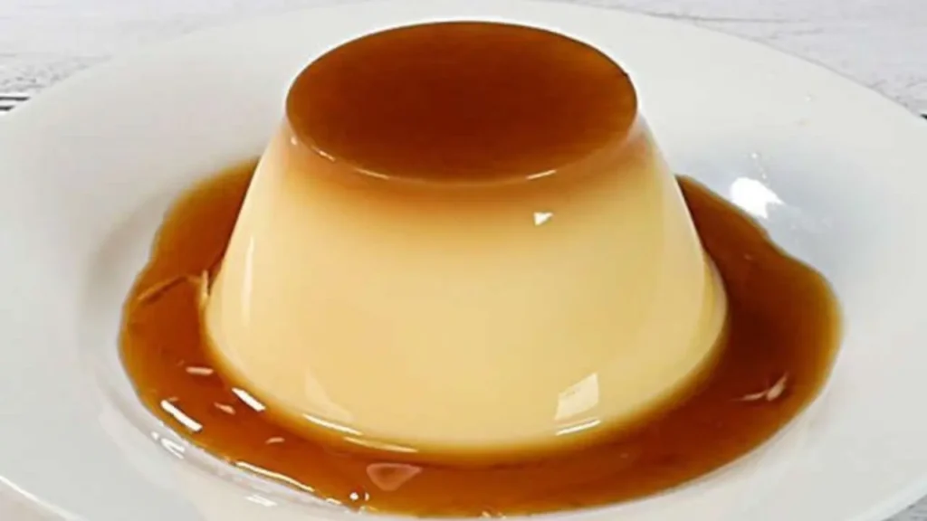 Pudim sem forno que fica cremoso, servido com calda de caramelo sobre prato branco.