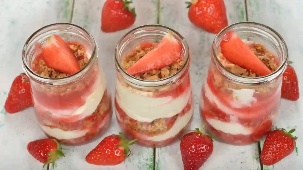 Três potes de vidro com camadas de creme, purê de morango e farofa crocante, decorados com morangos frescos — uma receita de sobremesa surpreendente e irresistível.