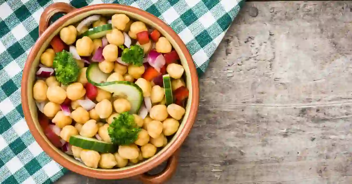 Salada de grão-de-bico com atum servida em bowl rústico, acompanhada de vegetais coloridos como pepino, pimentão vermelho, cebola roxa e brócolis, sobre superfície de madeira com pano xadrez verde e branco.