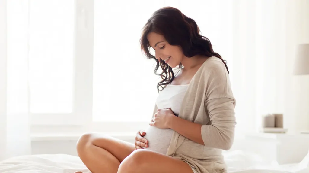 Mulher grávida sentada na cama, sorrindo e segurando a barriga com as mãos, em ambiente iluminado e tranquilo — representando os sintomas e estágios da gravidez.