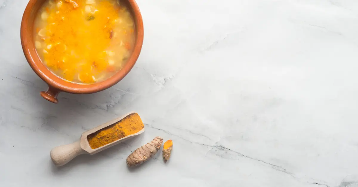 Tigela de sopa de frango com cúrcuma ao lado de uma colher de madeira com pó de cúrcuma e raiz fresca sobre superfície clara.