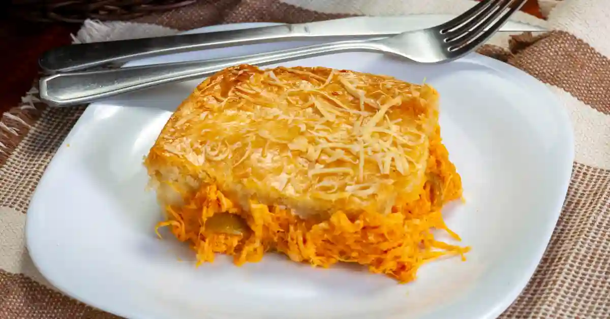Fatia de torta de frango com pão amanhecido servida em prato branco, coberta com queijo ralado.