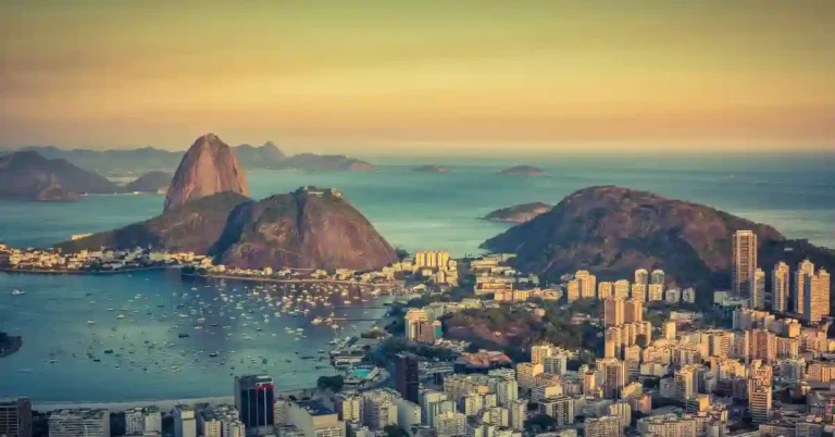 Vista panorâmica do Rio de Janeiro ao pôr do sol, com o Pão de Açúcar ao fundo e a cidade iluminada; cenário típico do trajeto do Trem do Corcovado.