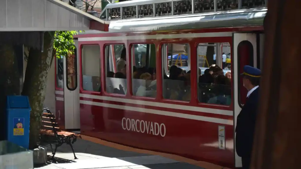 Trem do Corcovado vermelho com passageiros na estação, próximo a funcionário uniformizado e lixeira azul.