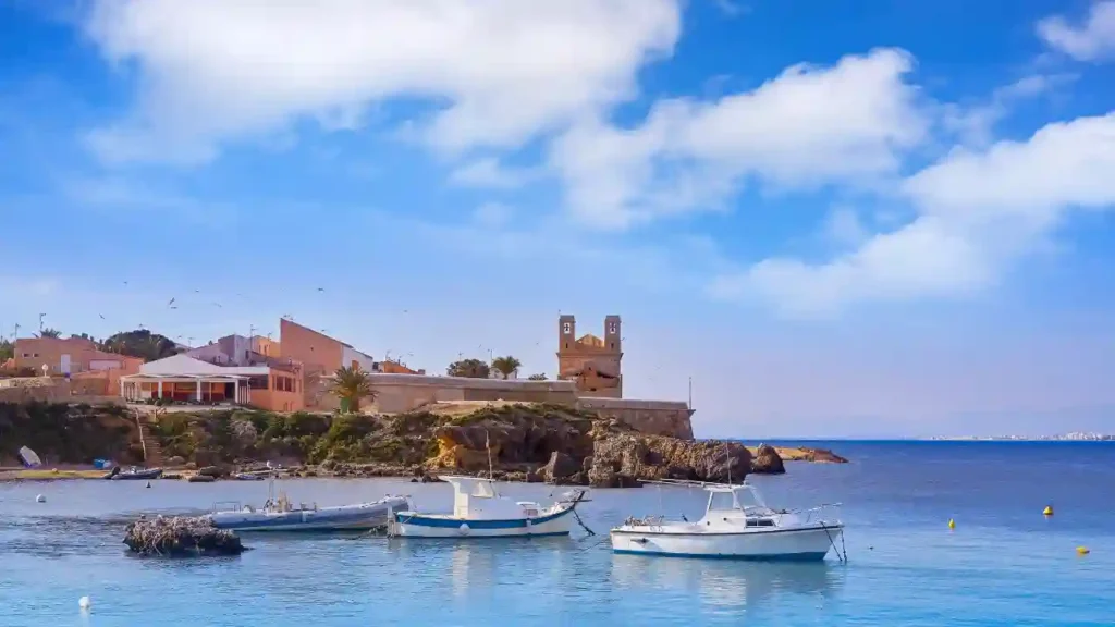 Três barcos ancorados em águas calmas diante de vilarejo à beira-mar com construções históricas e vegetação costeira sob céu azul.