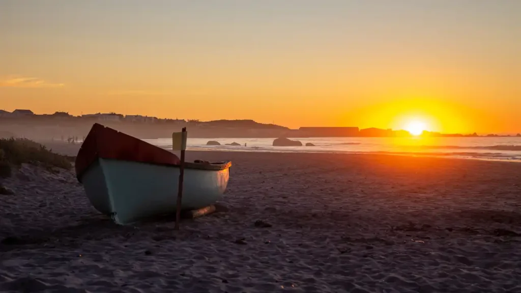 Barco na areia ao pôr do sol em praia tranquila, com vilarejo à beira-mar ao fundo e reflexos dourados sobre o mar.