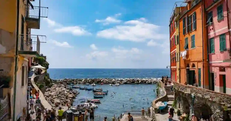 Vilarejo à beira-mar com construções coloridas, barcos ancorados e pessoas aproveitando o dia em um pequeno porto cercado pelo mar azul.