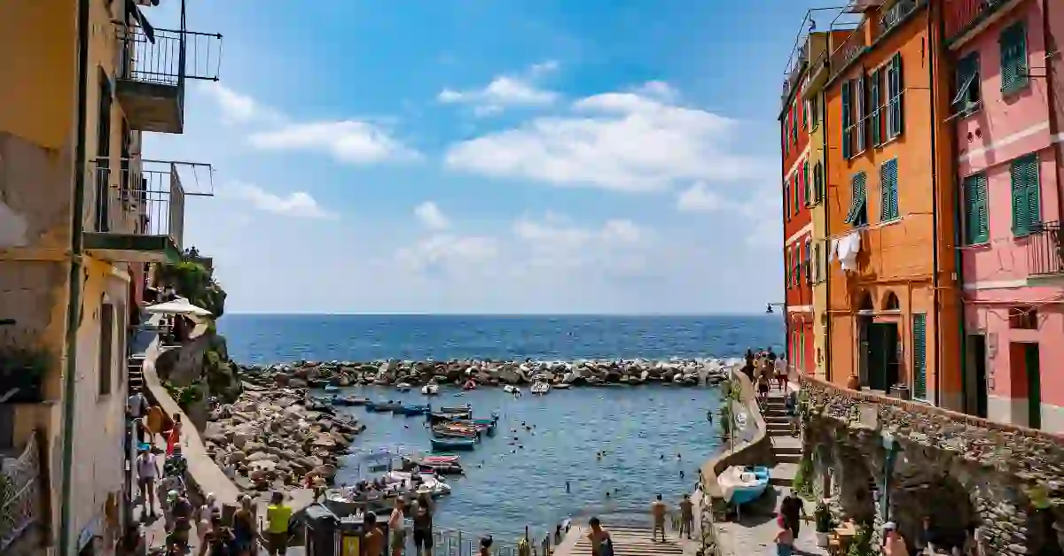 Vilarejo à beira-mar com construções coloridas, barcos ancorados e pessoas aproveitando o dia em um pequeno porto cercado pelo mar azul.