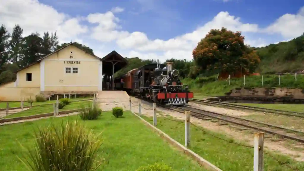 Locomotiva a vapor na estação Tiradentes, parte da ferrovia turística mais longa do Brasil, de Vitória a Minas, cercada por paisagem verde e céu parcialmente nublado.