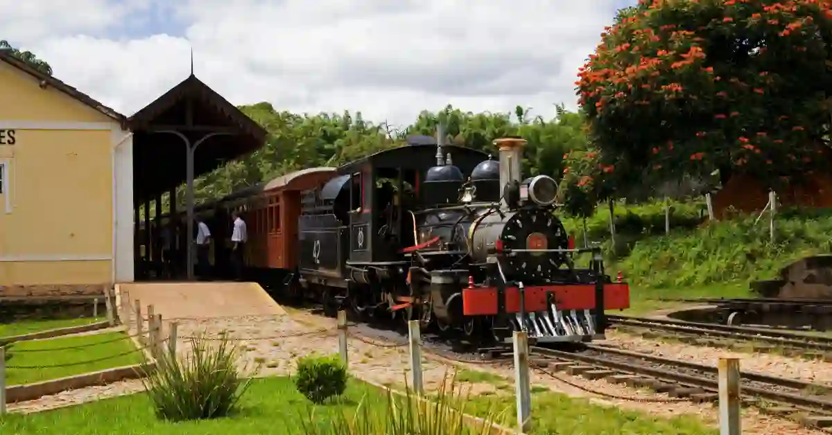 Locomotiva a vapor histórica na estação da ferrovia turística Vitória a Minas, a ferrovia turística mais longa do Brasil, cercada por vegetação e visitantes.
