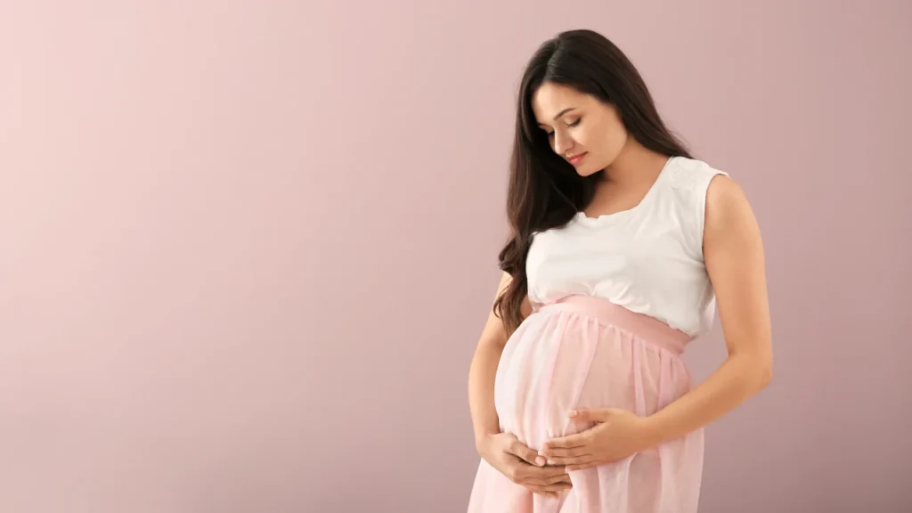 Você que está grávida, mulher acaricia a barriga com carinho, vestida com roupas claras, em frente a um fundo rosa suave.