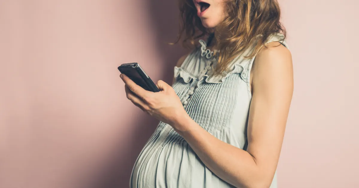 Pessoa grávida com expressão de surpresa ao olhar para o celular, diante de uma parede rosa — uma imagem que fala diretamente a você que está grávida.