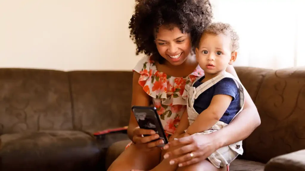 Mãe sorridente sentada no sofá com seu filho no colo, mostrando algo no celular, possivelmente um aplicativo de mães.