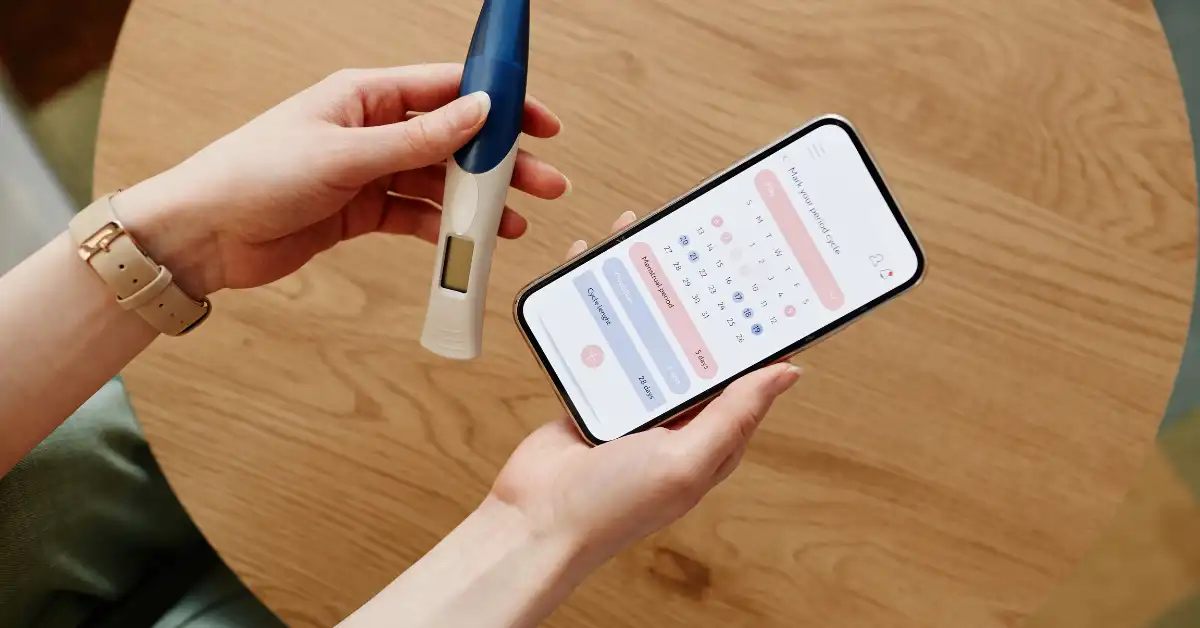 Pessoa segurando um termômetro digital e um smartphone com um aplicativo de mães voltado para o acompanhamento do ciclo menstrual.