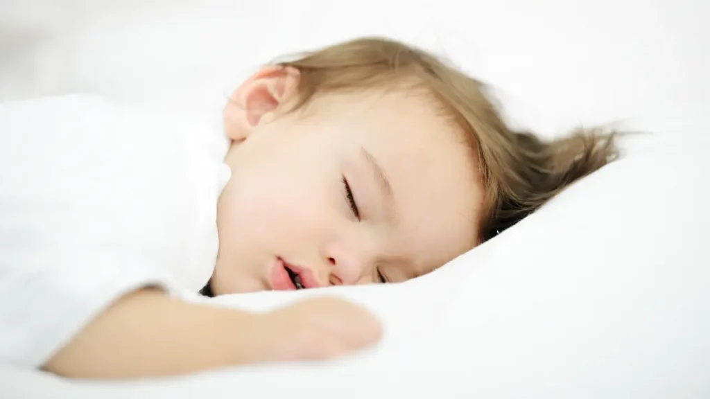 Criança dormindo serenamente em uma cama branca, representando os efeitos calmantes de um aplicativo de sono bebê.