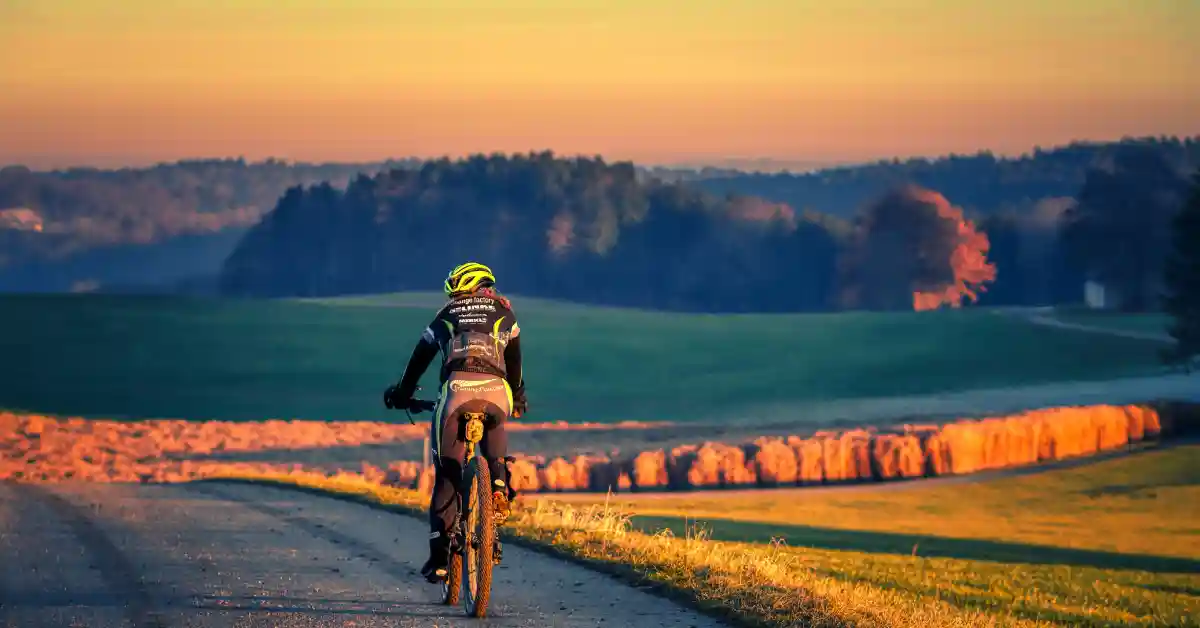 Ciclista pedalando por estrada cercada por campos e florestas ao pôr do sol, evocando a experiência do ciclismo na Patagônia.