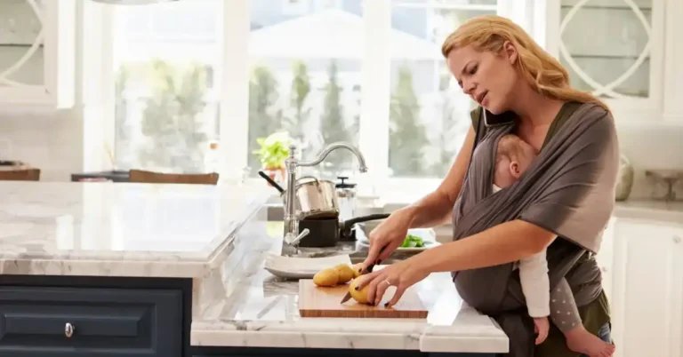 Mulher na cozinha cortando batatas enquanto segura o bebê em um sling e fala ao telefone — uma cena que retrata sua experiência como mãe.