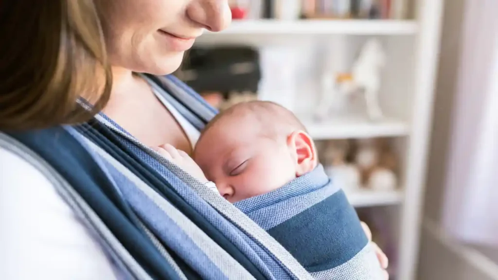 Mulher sorridente segura seu bebê adormecido em um sling azul listrado, representando um momento íntimo da experiência como mãe.