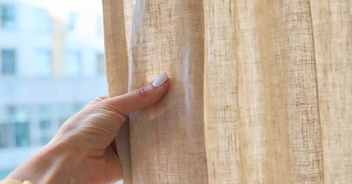 Mão com esmalte claro segurando uma cortina bege texturizada, com fundo desfocado de janelas ao fundo — ideal para ilustrar dicas de como limpar cortina sem tirar.