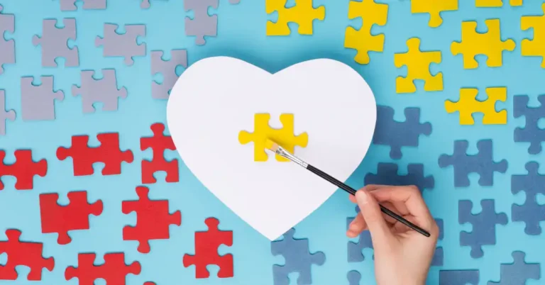 Mão pintando peça de quebra-cabeça amarela sobre recorte em forma de coração, cercado por peças coloridas, representando apoio e criatividade das mães de neurodivergentes.