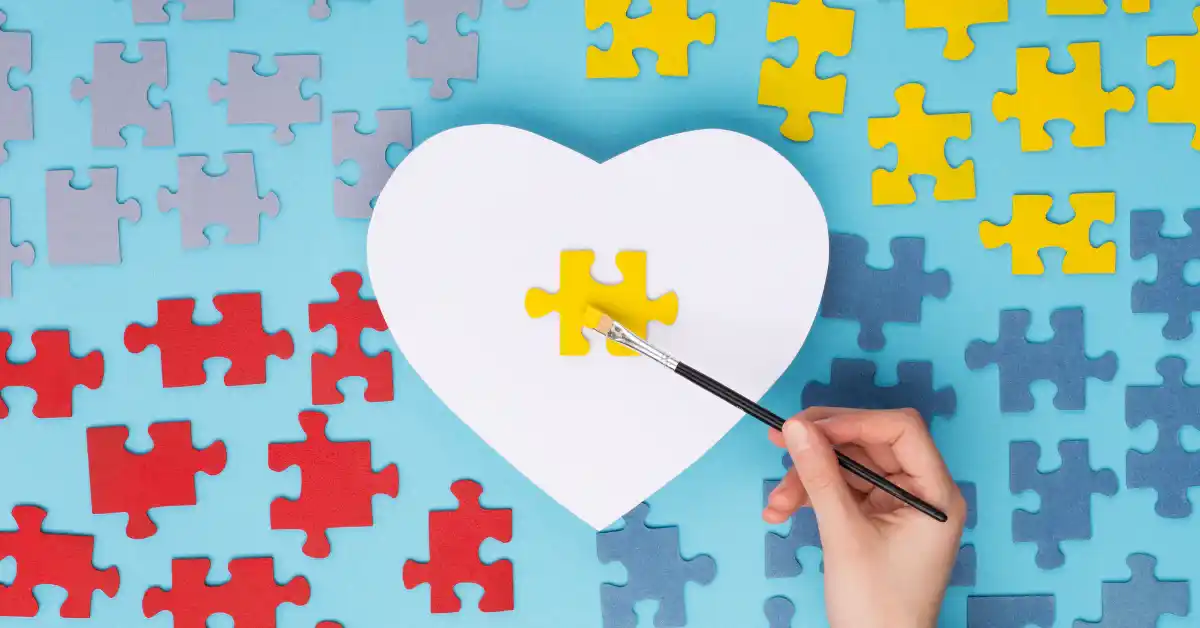 Mão pintando peça de quebra-cabeça amarela sobre recorte em forma de coração, cercado por peças coloridas, representando apoio e criatividade das mães de neurodivergentes.