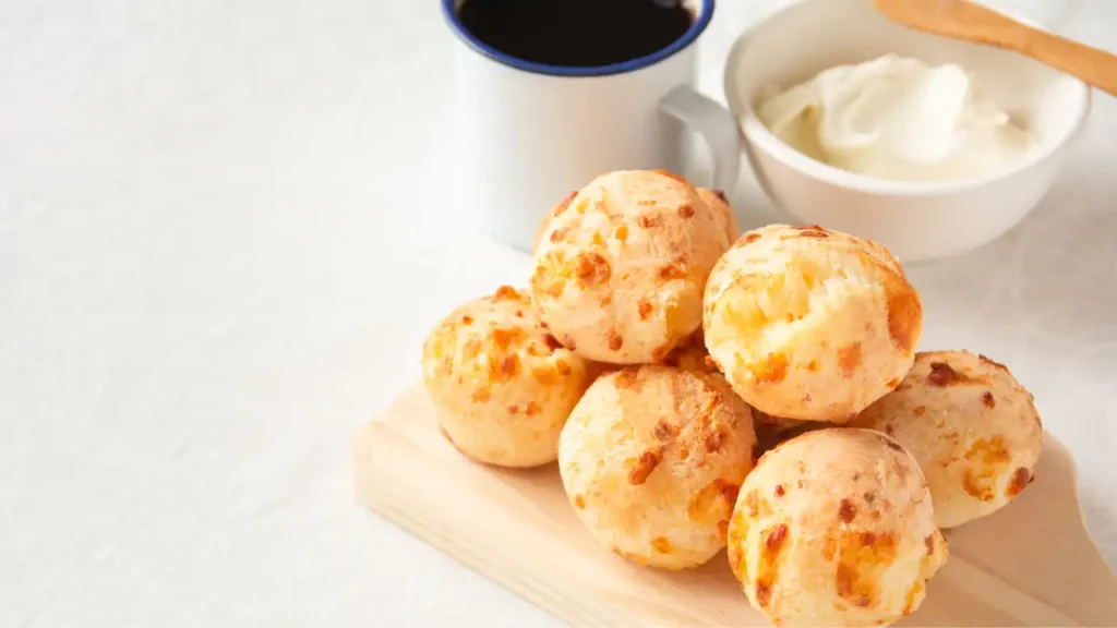Pão de queijo crocante servido sobre tábua de madeira, acompanhado de café preto e creme branco em tigela.