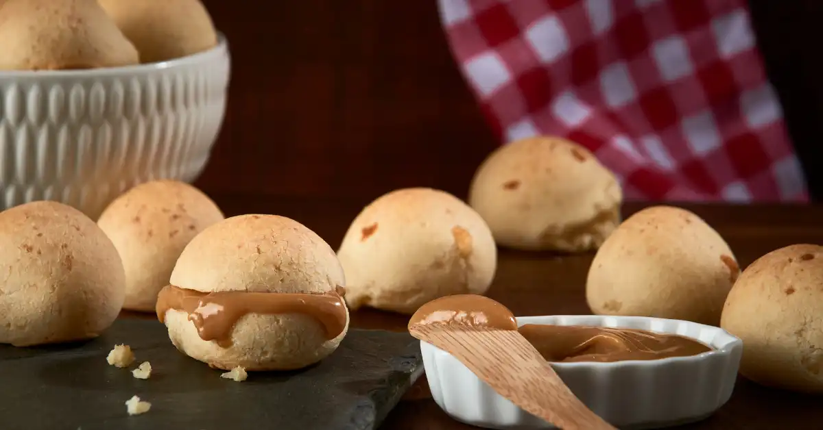 Pão de queijo crocante recheado com doce de leite, servido com creme extra em prato branco e pano xadrez ao fundo.