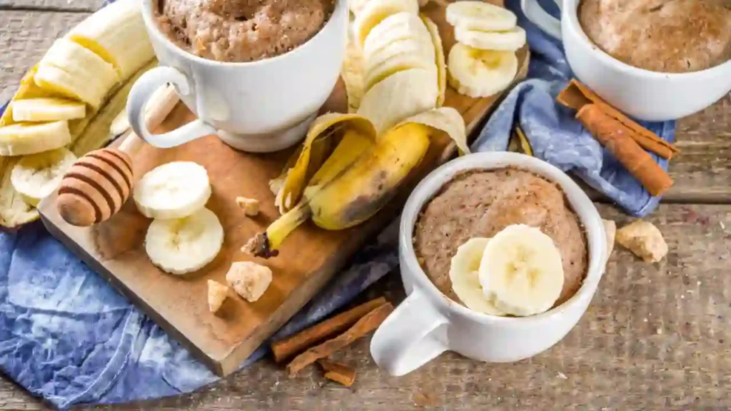 Três canecas brancas com bolo de banana servido sobre uma mesa rústica, cercadas por fatias de banana, mel, açúcar mascavo e canela — uma sugestão acolhedora para receitas de lanche fáceis e rápidas.