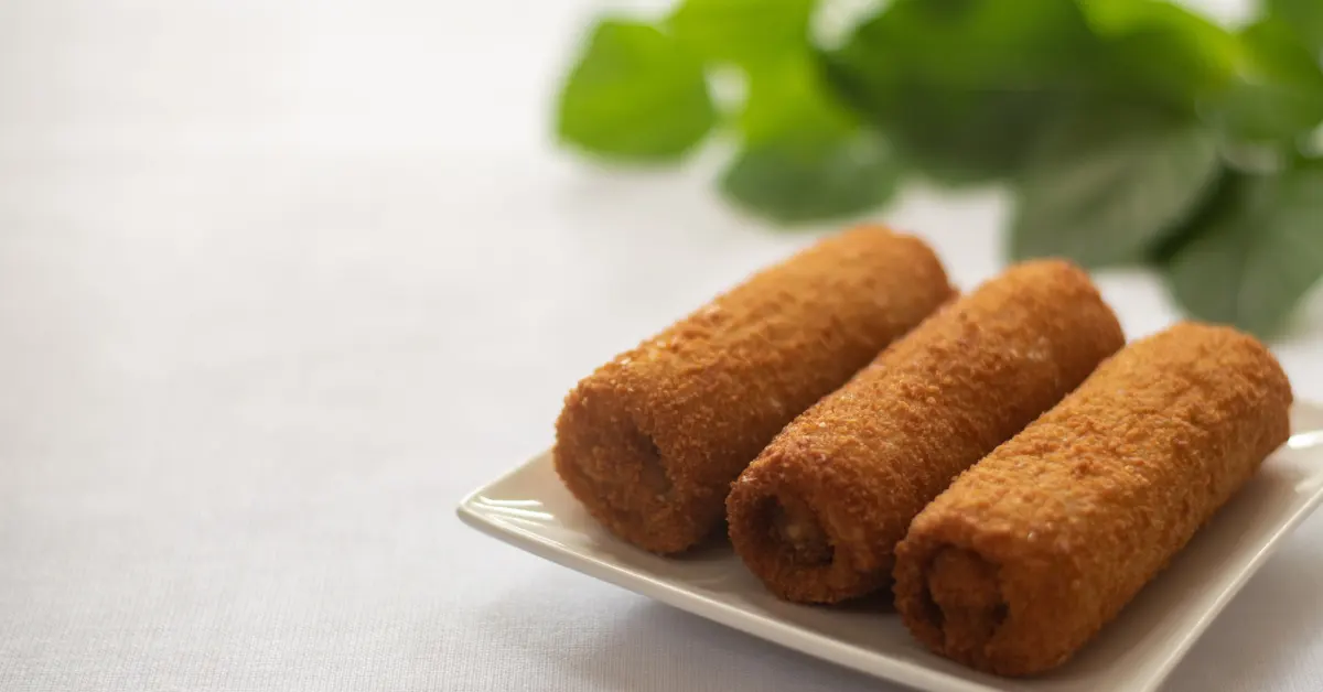 Três croquetes dourados e empanados servidos em um prato branco, com folhas verdes desfocadas ao fundo — uma sugestão deliciosa para receitas de lanche fáceis e rápidas.