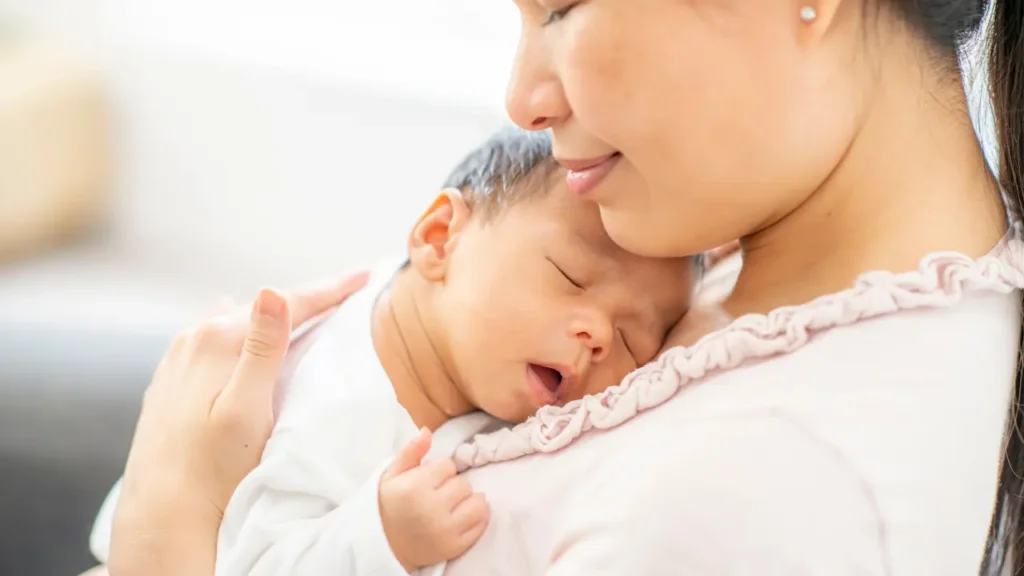Mãe segurando seu bebê recém-nascido adormecido no colo, representando o vínculo nos primeiros meses de vida.